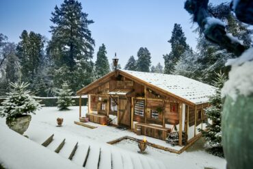 Auf einer verschneiten Gartenterrasse steht eine temporär aufgebaute Almhütte aus Holz. Alles ist verschneit. Rund um die Terrasse, zu der eine Treppe hinabführt, zieht sich eine gepflegte Hecke, dahinter rundum mehrere Bäume.