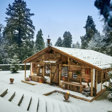 Auf einer verschneiten Gartenterrasse steht eine temporär aufgebaute Almhütte aus Holz. Alles ist verschneit. Rund um die Terrasse, zu der eine Treppe hinabführt, zieht sich eine gepflegte Hecke, dahinter rundum mehrere Bäume.