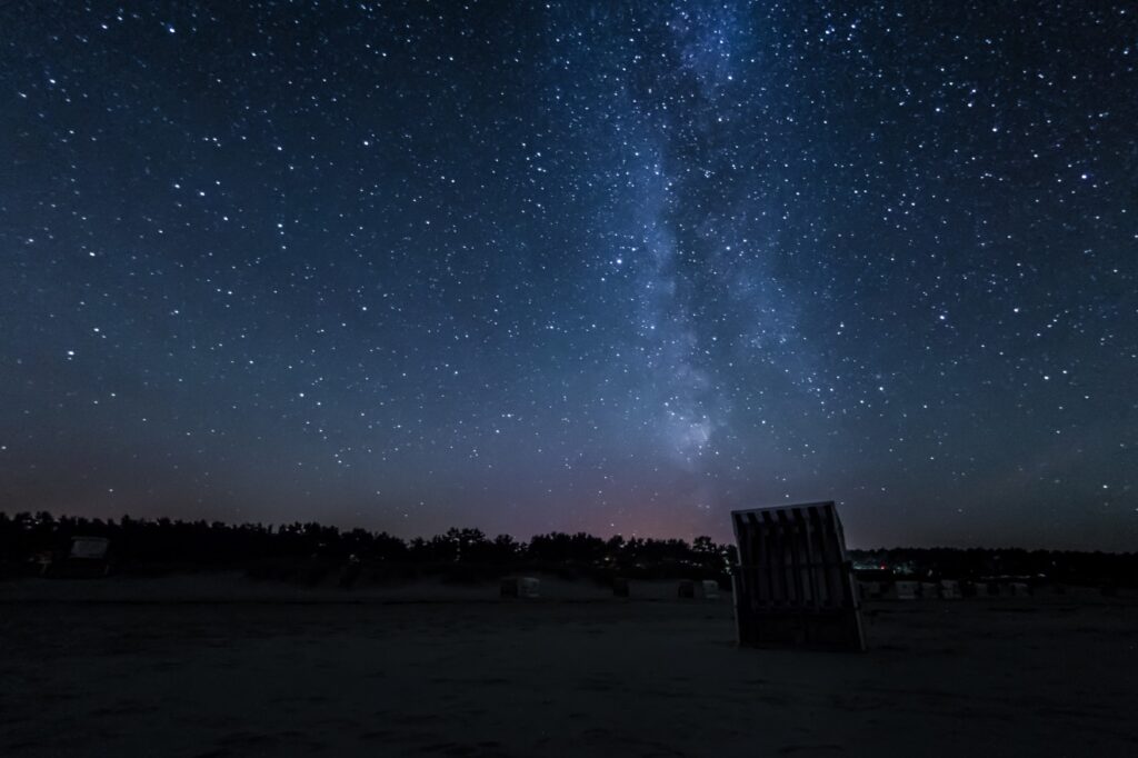 Ein klarer Sternenhimmel mit zahllosen Sternen. Im rechten Drittel scheint die Milchstraße. Am unteren Bildrand ist die Silhouette eines Strandkorbs erkennbar.
