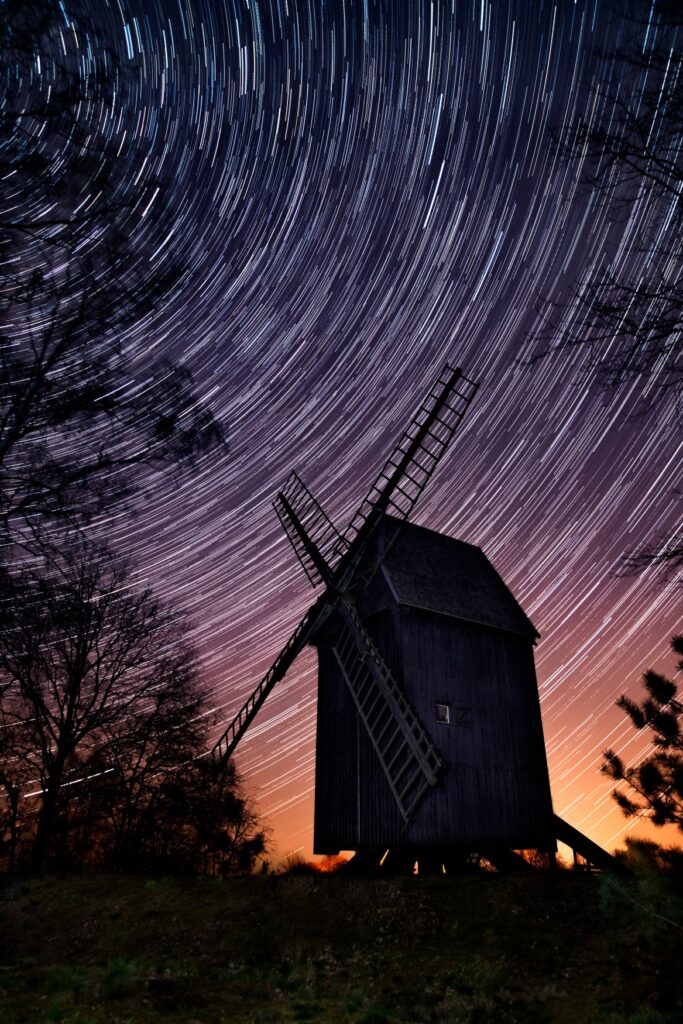Vor der Langzeitbelichtung eines Nachthimmels zeichnet sich die dunkle Silhouette einer historischen Windmühle ab. Die Sterne bilden eine Art "Scheibe", der untere Rand des Himmels leuchtet noch leicht gelb.