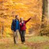 Ein junger Mann und eine junge Frau in bunter Outdoorkleidung laufen auf einem Waldweg in Richtung des Betrachters. Beide schauen nach rechts, die Frau zeigt auf etwas. Der Wald um sie herum ist vollständig in Rot- und Gelbtönen.