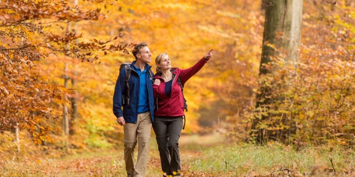 Ein junger Mann und eine junge Frau in bunter Outdoorkleidung laufen auf einem Waldweg in Richtung des Betrachters. Beide schauen nach rechts, die Frau zeigt auf etwas. Der Wald um sie herum ist vollständig in Rot- und Gelbtönen.