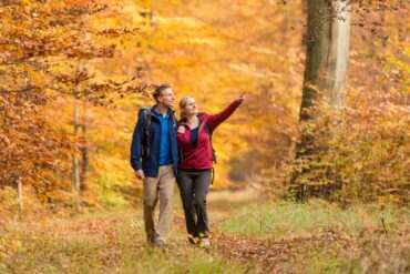 Ein junger Mann und eine junge Frau in bunter Outdoorkleidung laufen auf einem Waldweg in Richtung des Betrachters. Beide schauen nach rechts, die Frau zeigt auf etwas. Der Wald um sie herum ist vollständig in Rot- und Gelbtönen.