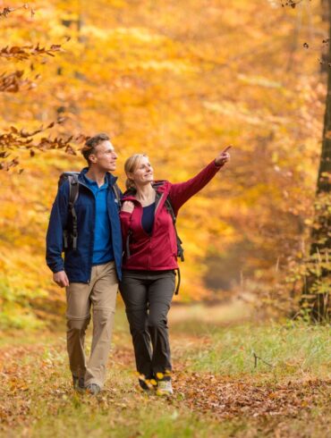 Ein junger Mann und eine junge Frau in bunter Outdoorkleidung laufen auf einem Waldweg in Richtung des Betrachters. Beide schauen nach rechts, die Frau zeigt auf etwas. Der Wald um sie herum ist vollständig in Rot- und Gelbtönen.