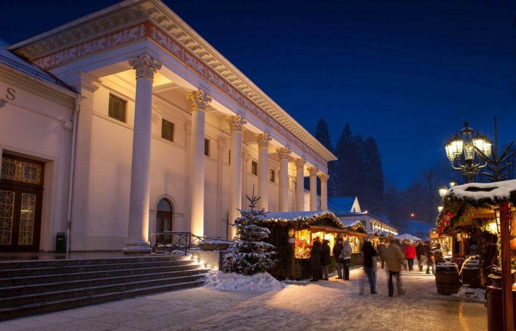 Vor einem weißen, klassizistischen Palas zieht sich die hell erleuchtete Gasse eines Weihnachtsmarkts entlang. Der Himmel ist dunkel, der Boden mit Schnee bedeckt.