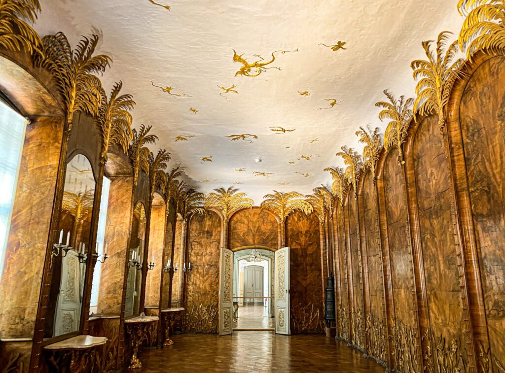 A magnificent hall in a castle. The walls are panelled with fine wood, and carved palm trees are placed between the individual sections, their leaves made of golden plates on the ceiling. The floor is parquet, and sunlight shines through the windows on the left side of the hall. Right in the centre of the picture, a double door opens into the next room.