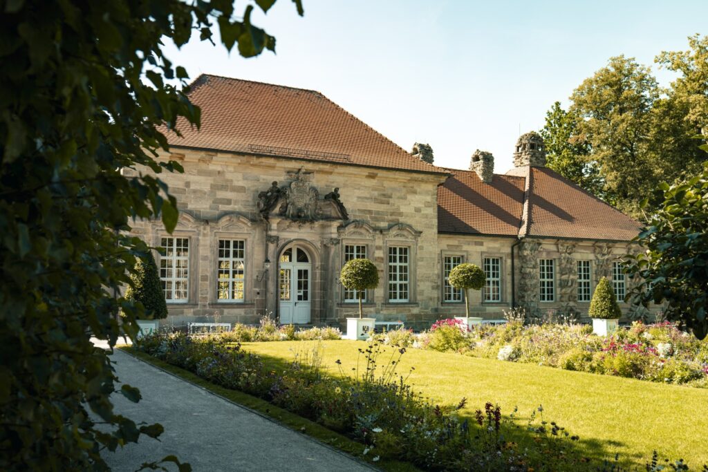 A low Baroque building made of gray stone blocks with tall, white windows on the ground floor and a red tile roof. In front of it lies a small, well-kept garden.