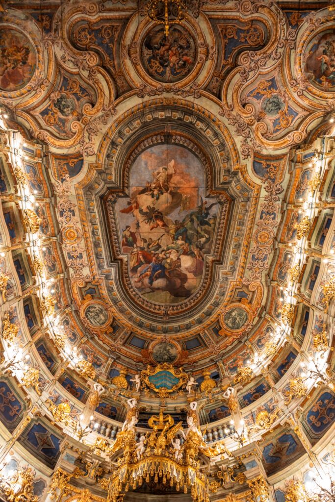 Ceiling fresco in the Margravial Opera House in Bayreuth; in the center of the fresco is a scene depicting several people sitting on a kind of cloud tower and playing harps; the audience seating is arranged in tiers around the perimeter. Everything is brightly lit and colorfully painted.