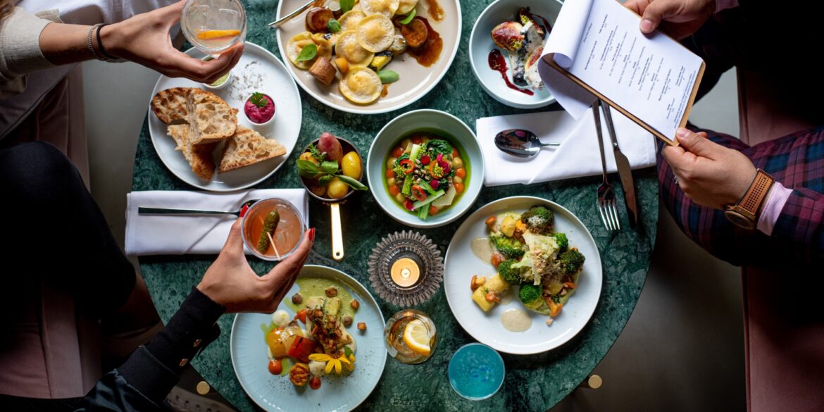 Ein runder Tisch aus grünem Marmor, der rappelvoll mit kleinen und großen Speisen beladen ist. Drei Personen sitzen am Tisch, auf dem Bild sind nur ihre Beine von oben zu sehen sowie die Getränke der beiden Personen zur Linken, die diese über den Tisch halten. Die Person rechts hingegen hält die Speisekarte in Händen.