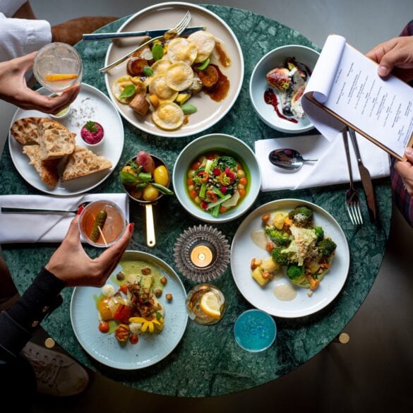 Ein runder Tisch aus grünem Marmor, der rappelvoll mit kleinen und großen Speisen beladen ist. Drei Personen sitzen am Tisch, auf dem Bild sind nur ihre Beine von oben zu sehen sowie die Getränke der beiden Personen zur Linken, die diese über den Tisch halten. Die Person rechts hingegen hält die Speisekarte in Händen.