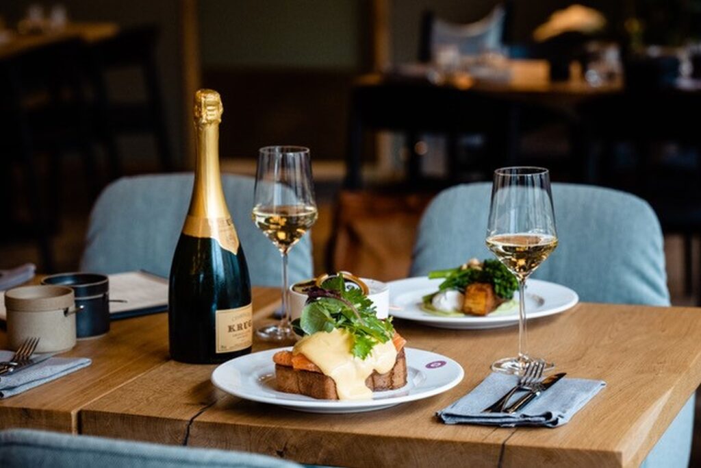 Auf einem Holztisch in einem Restaurant steht ein Teller mit einem großen Croque Madame, daneben eine Flasche Cremant mit zwei Gläsern. Die Polsterstühle hinter dem Tisch sind hellblau.