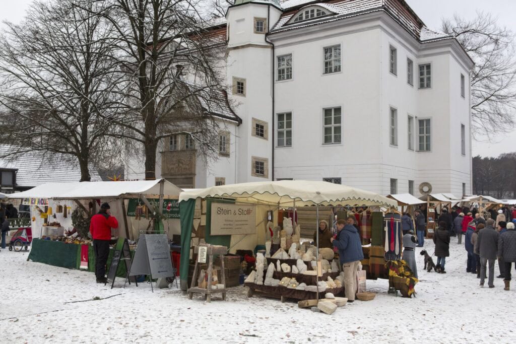 A small christmas market in front of a small historic castle. Everthing is covered in snow.