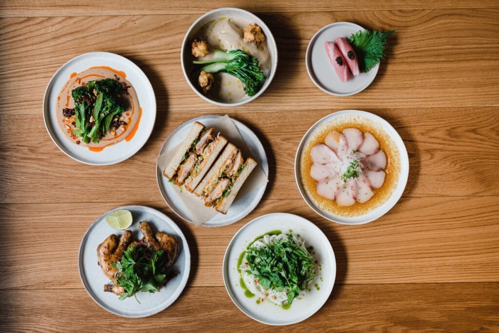 Auf einem Holztisch stehen kreisrund angeordnet sieben kunstvoll arrangierte Teller mit verschiedenen Gerichten. Alle sind so portioniert, dass man sie gut teilen kann.