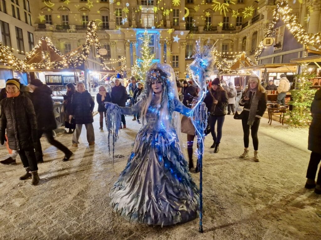 A person in a high quality, blue ice sorceress costume stands on a brightly lighted christmas market.