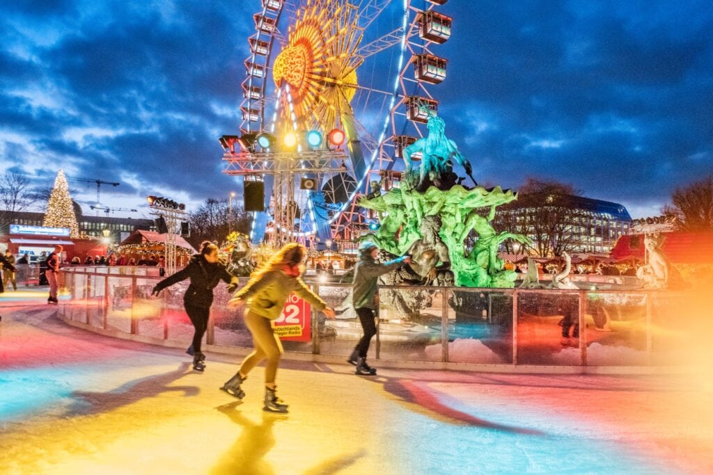 People skating on an ice rink that leads around a central place with a giant ferris wheel. Everything is colorfully illuminated.