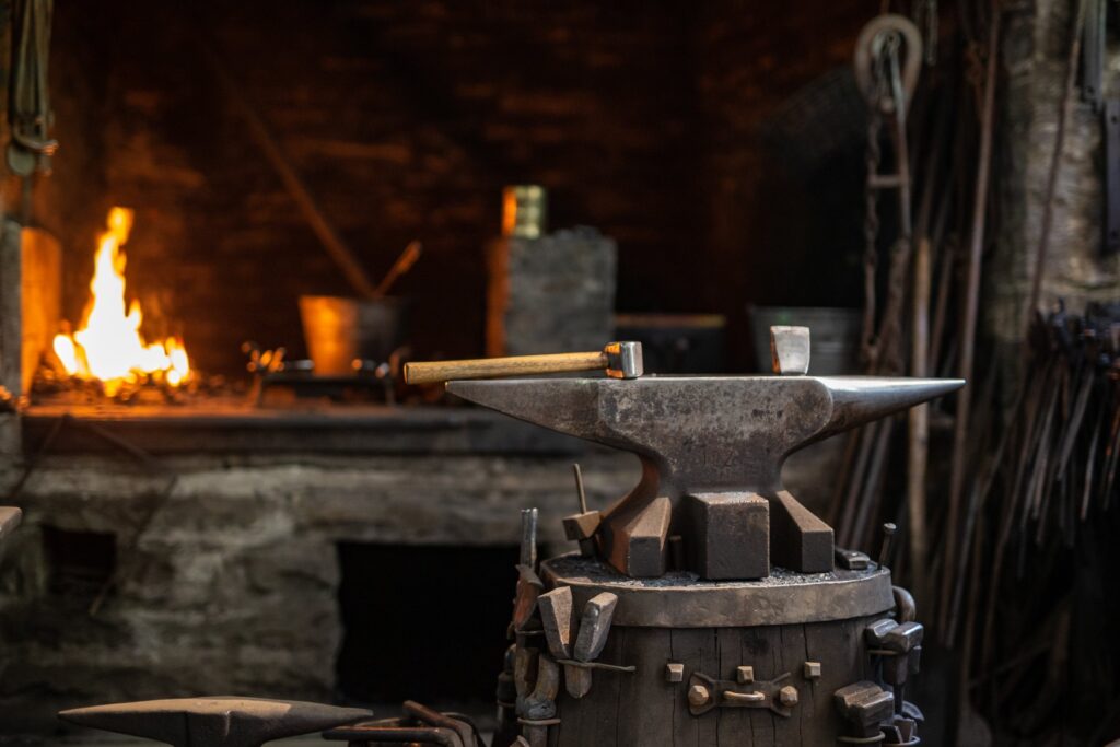 Ein Amboss steht in einer Schmiede auf einem Holzfass, darauf liegen ein Hammer und ein STück Metall. Am Holzfass sind mehrere Metallteile angebracht. Im Hintergrund ist links unscharf eine Esse zu erkennen, rechts lehnen mehrere Metallstäbe an einer Steinsäule.
