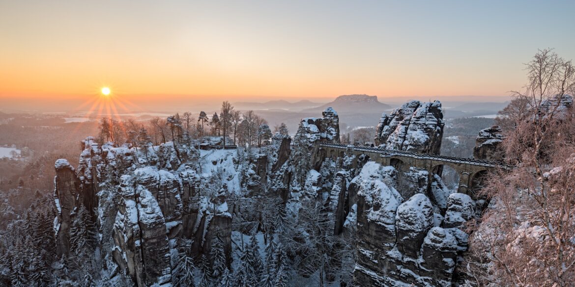 Über ein verschneites Felsenmeer spannt eine mächtige Sandsteinbrücke vom rechten Bildrand bis zu einem mittigen Felsplateau. Dahinter eine weite Ebene, über der gerade die Wintersonne an einem klaren Himmel untergeht.
