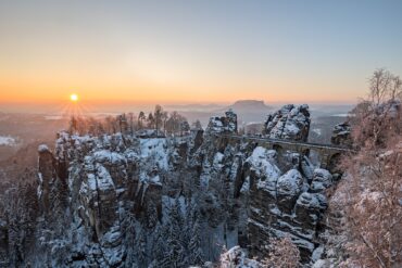 Über ein verschneites Felsenmeer spannt eine mächtige Sandsteinbrücke vom rechten Bildrand bis zu einem mittigen Felsplateau. Dahinter eine weite Ebene, über der gerade die Wintersonne an einem klaren Himmel untergeht.