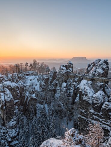 Über ein verschneites Felsenmeer spannt eine mächtige Sandsteinbrücke vom rechten Bildrand bis zu einem mittigen Felsplateau. Dahinter eine weite Ebene, über der gerade die Wintersonne an einem klaren Himmel untergeht.