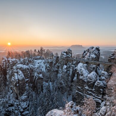 Über ein verschneites Felsenmeer spannt eine mächtige Sandsteinbrücke vom rechten Bildrand bis zu einem mittigen Felsplateau. Dahinter eine weite Ebene, über der gerade die Wintersonne an einem klaren Himmel untergeht.