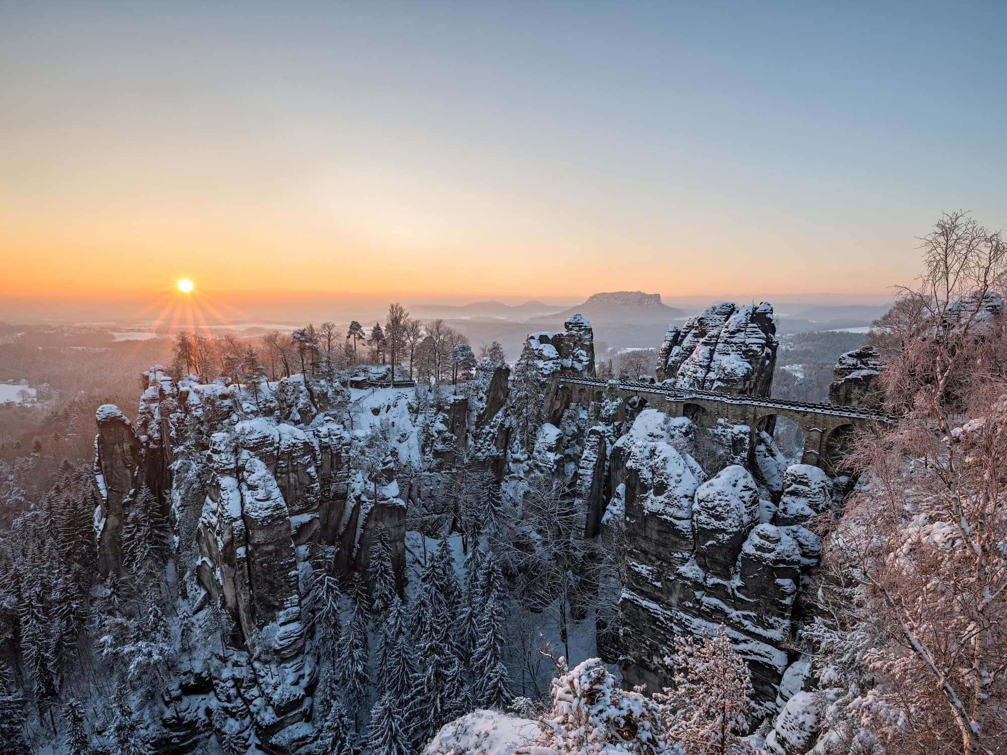 Über ein verschneites Felsenmeer spannt eine mächtige Sandsteinbrücke vom rechten Bildrand bis zu einem mittigen Felsplateau. Dahinter eine weite Ebene, über der gerade die Wintersonne an einem klaren Himmel untergeht.