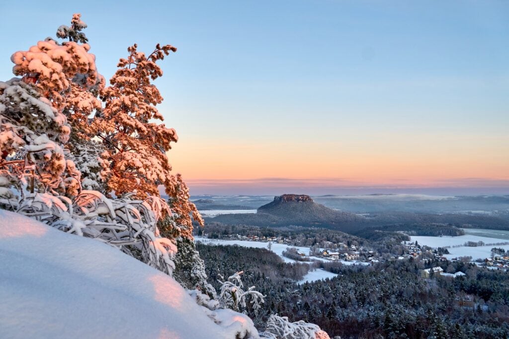 Zwei dick verschneite Nadelbäume stehen auf der linken auf einem verschneiten Hang. Dahinter öffnet sich ein weiter Blick in ein Tal, teilweise verschneit und mit einigen Dörfern. Am Horizont zeigt sich schon ein roter Streifen des Sonnenuntergangs, darüber ist der Himmel noch blau.