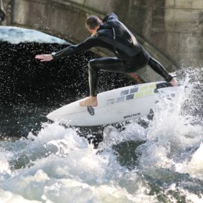 A man in a wetsuit is pushed up by a wave on his surfboard. He has his arms outstretched on both sides.