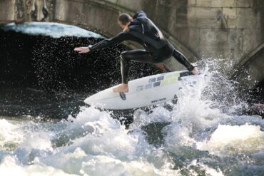 A man in a wetsuit is pushed up by a wave on his surfboard. He has his arms outstretched on both sides.