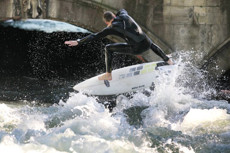 A man in a wetsuit is pushed up by a wave on his surfboard. He has his arms outstretched on both sides.