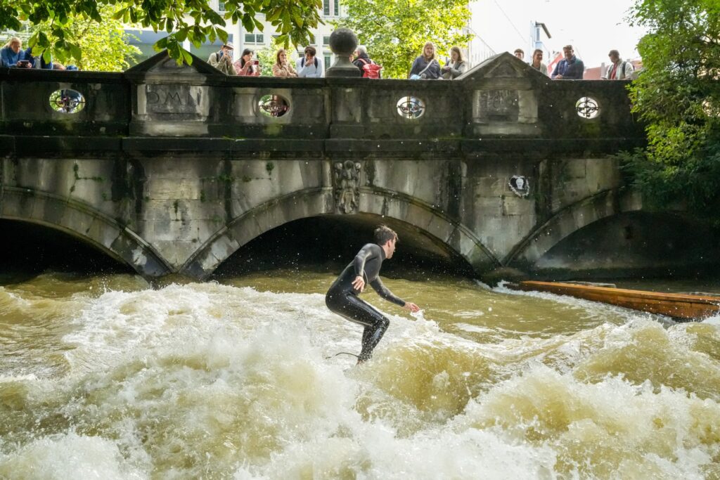 A man in a wetsuit surfs on a standing wave in a river. The river flows out from under a bridge. People on the bridge watch the activity on the river.
