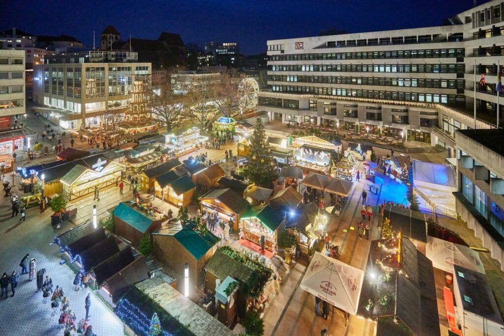 Drohnenansicht eines Weihnachtsmarkts auf einem großen Platz, alles ist hell erleuchtet, im Hintergrund ist ein Riesenrad zu sehen.