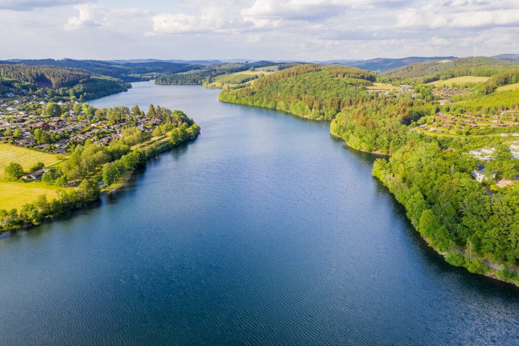 Ein langezogener, tiefblauer See, zu beiden Seiten von waldigem Ufer begrenzt, aufgenommen mit einer Drohne. Auf einer Landzunge auf der linken Seite ist eine kleine Stadt.