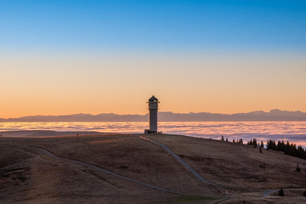 Auf einer kahlen Hügelkuppe steht einsam ein hoher Aussichtsturm. Das Tal dahinter ist wolkenverhangen, am Horizont zeichnen sich im Sonnenuntergang die Alpen ab.