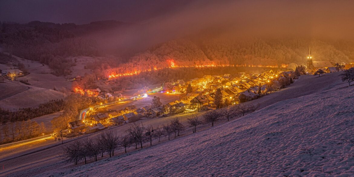 Blick von einer Anhöhe auf eine zur Weihnachtszeit hell erleuchtete Kleinstadt im Tal. Hinter der Stadt führt ein hell beleuchteter Pfad durch den Wald. Alles liegt unter einer dicken Schneedecke.
