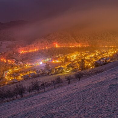 Blick von einer Anhöhe auf eine zur Weihnachtszeit hell erleuchtete Kleinstadt im Tal. Hinter der Stadt führt ein hell beleuchteter Pfad durch den Wald. Alles liegt unter einer dicken Schneedecke.