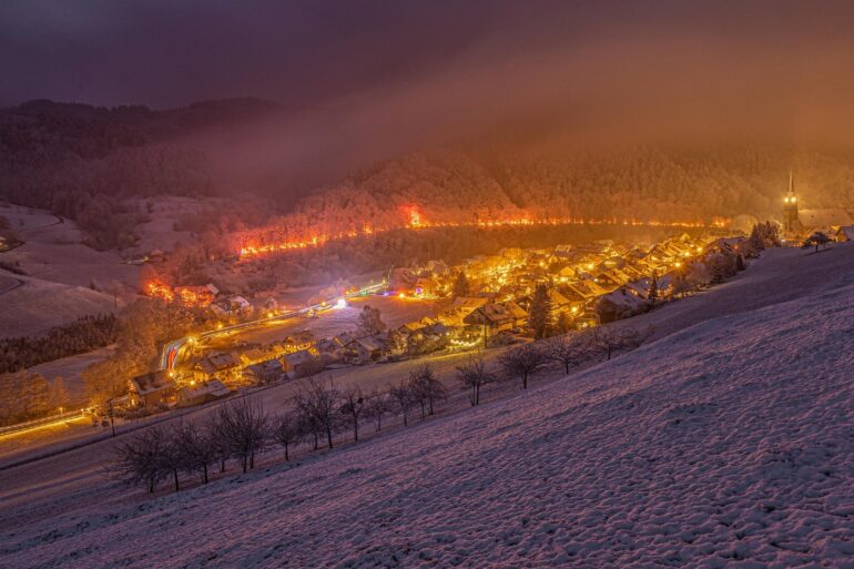 Blick von einer Anhöhe auf eine zur Weihnachtszeit hell erleuchtete Kleinstadt im Tal. Hinter der Stadt führt ein hell beleuchteter Pfad durch den Wald. Alles liegt unter einer dicken Schneedecke.