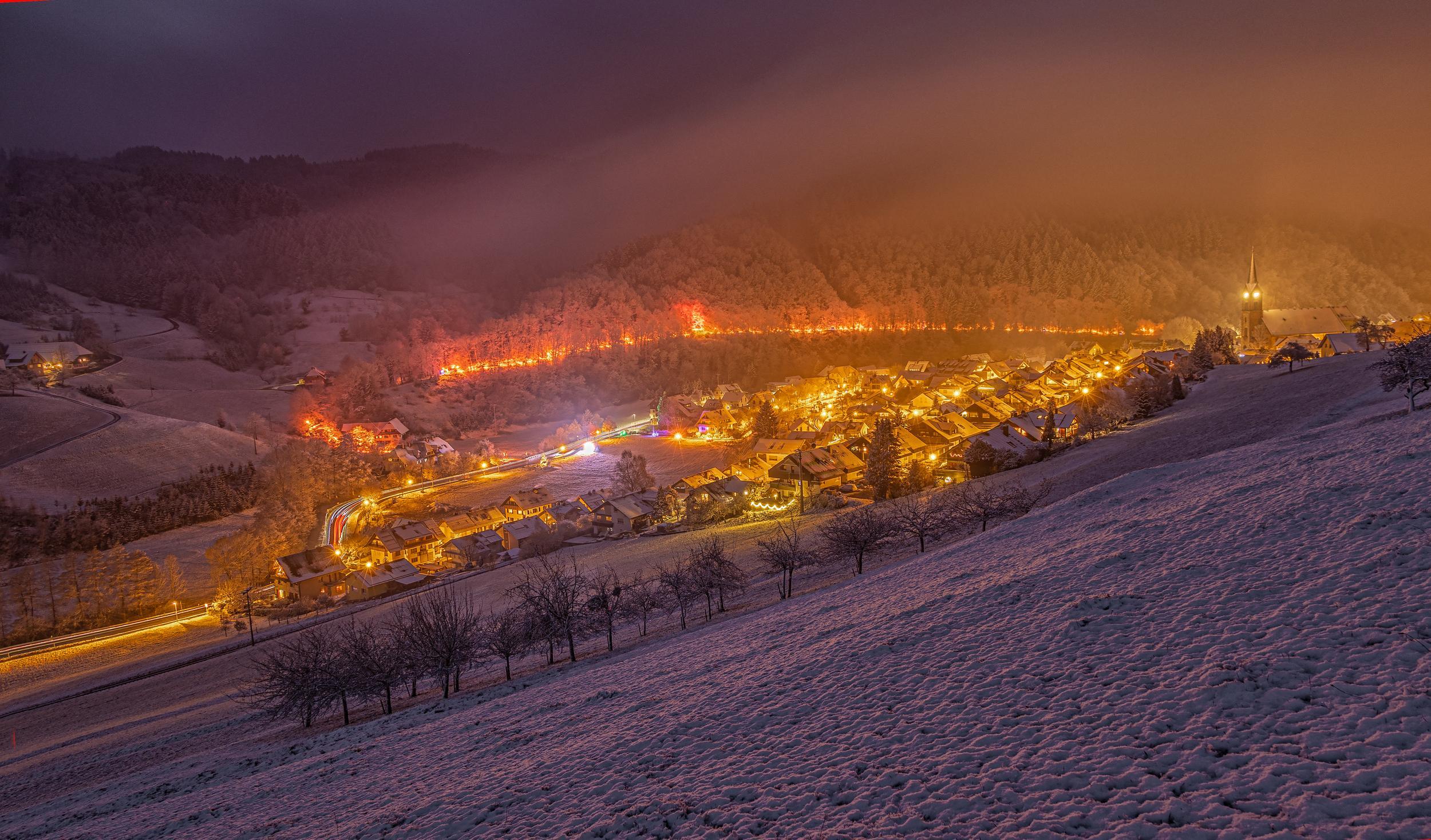 Blick von einer Anhöhe auf eine zur Weihnachtszeit hell erleuchtete Kleinstadt im Tal. Hinter der Stadt führt ein hell beleuchteter Pfad durch den Wald. Alles liegt unter einer dicken Schneedecke.