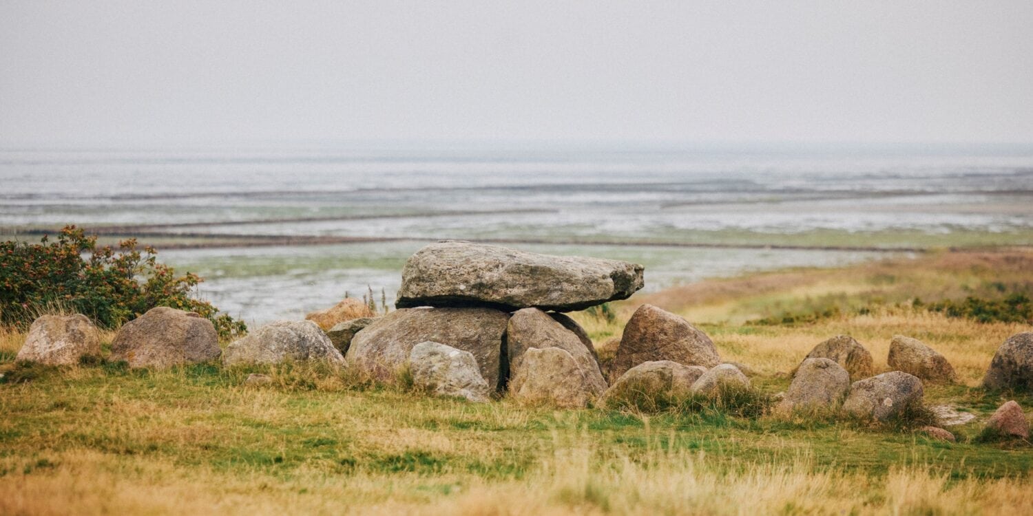 Auf einer herbstlichen Düne steht ein Hünengrab, eine Ansammlung großer Steine, über die quer eine Steinplatte gelegt wurde. Im Hintergrund geht eine Salzwiese ins Watt über, darüber diesig-grauer Himmel.