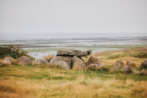 Auf einer herbstlichen Düne steht ein Hünengrab, eine Ansammlung großer Steine, über die quer eine Steinplatte gelegt wurde. Im Hintergrund geht eine Salzwiese ins Watt über, darüber diesig-grauer Himmel.