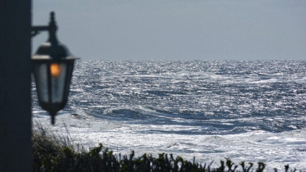Die wilde Nordsee im winterlichen Sonnenlicht. Hohe Wellen schlagen übereinander. Im Vordergrund hängt auf der linken Bildseite unscharf eine Metalllaterne, am unteren Bildrand unscharf etwas Strandbewuchs.
