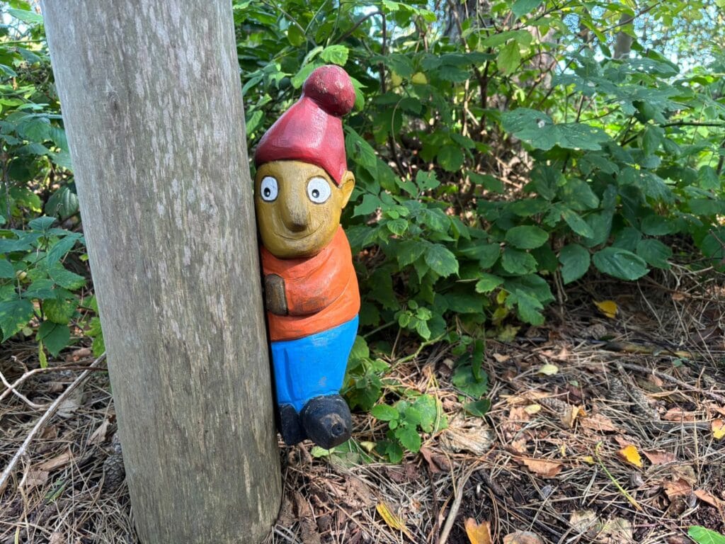 Hinter einem Holzpfahl lugt eine kleine Figur hervor, die wie ein Zwerg oder Wichtel gekleidet ist, mit roter Zipfelmütze.