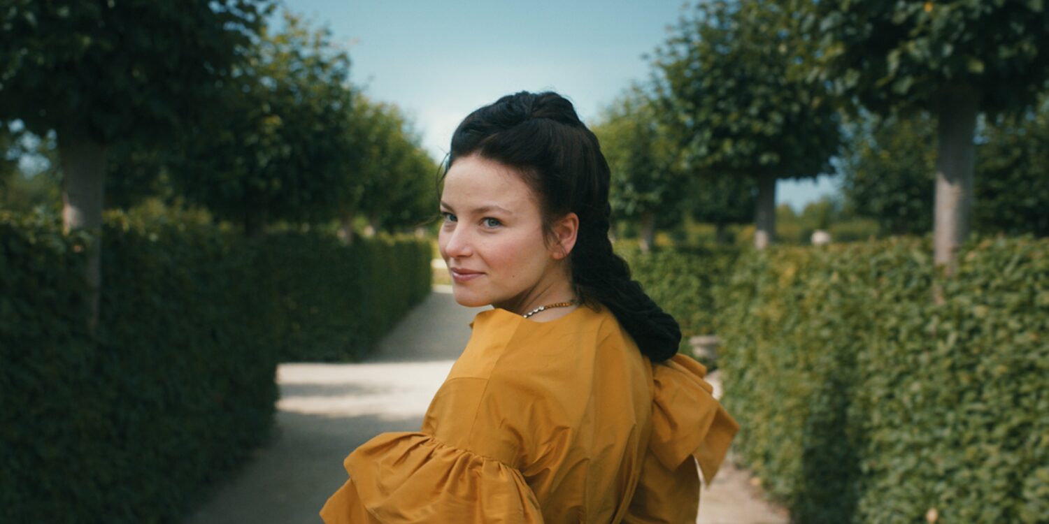 A young woman in a magnificent yellow dress with dark hair tied back in an elaborate braid stands with her back to the viewer in a well-tended garden, looking over her shoulder to the left. On both sides, tall hedges stretch toward a vanishing point hidden behind the woman.