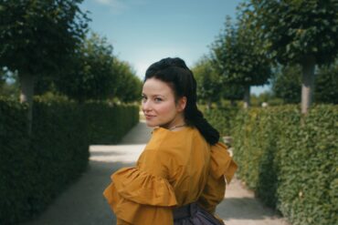 A young woman in a magnificent yellow dress with dark hair tied back in an elaborate braid stands with her back to the viewer in a well-tended garden, looking over her shoulder to the left. On both sides, tall hedges stretch toward a vanishing point hidden behind the woman.