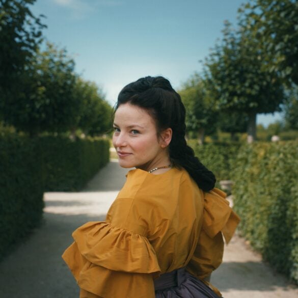 A young woman in a magnificent yellow dress with dark hair tied back in an elaborate braid stands with her back to the viewer in a well-tended garden, looking over her shoulder to the left. On both sides, tall hedges stretch toward a vanishing point hidden behind the woman.