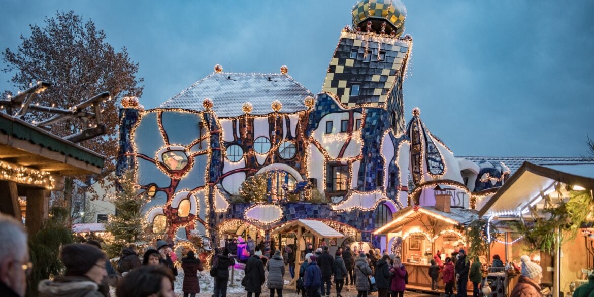 Auf einem gepflasterten Platz stehen mehrere beleuchtete Hütten eines Weihnachtsmarkts, der von vielen Menschen besucht ist. Im Hintergrund ist eine hohe Halle mit einem Turm des Architekten Hundertwasser; das ganze Gebäude scheint ineinander verdreht zu sein.