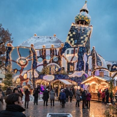 Auf einem gepflasterten Platz stehen mehrere beleuchtete Hütten eines Weihnachtsmarkts, der von vielen Menschen besucht ist. Im Hintergrund ist eine hohe Halle mit einem Turm des Architekten Hundertwasser; das ganze Gebäude scheint ineinander verdreht zu sein.