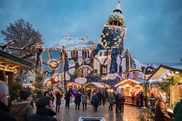 Auf einem gepflasterten Platz stehen mehrere beleuchtete Hütten eines Weihnachtsmarkts, der von vielen Menschen besucht ist. Im Hintergrund ist eine hohe Halle mit einem Turm des Architekten Hundertwasser; das ganze Gebäude scheint ineinander verdreht zu sein.
