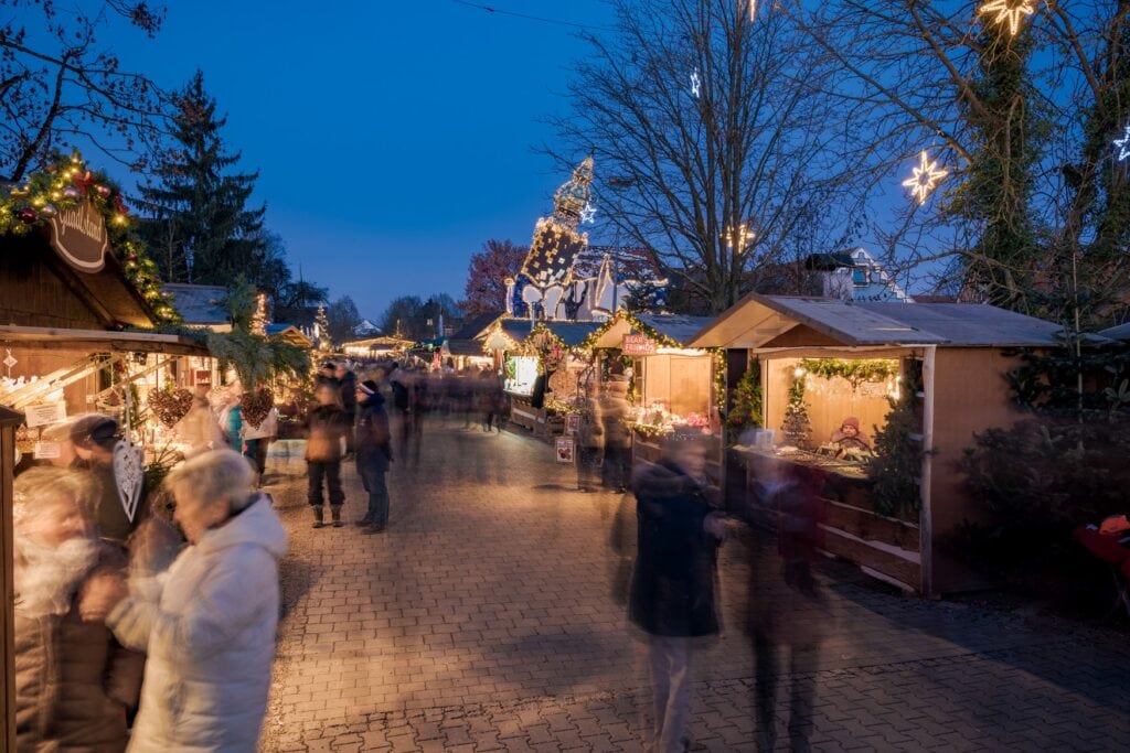 Eine "Gasse" auf einem Weihnachtsmarkt, auf der viele Menschen unterwegs sind. Durch eine Langzeitbelichtung sind die Personen unscharf und "verschwimmen" ineinander. Im Hintergrund ist ein schief gebauter Turm des Architekten Hundertwasser zu sehen.