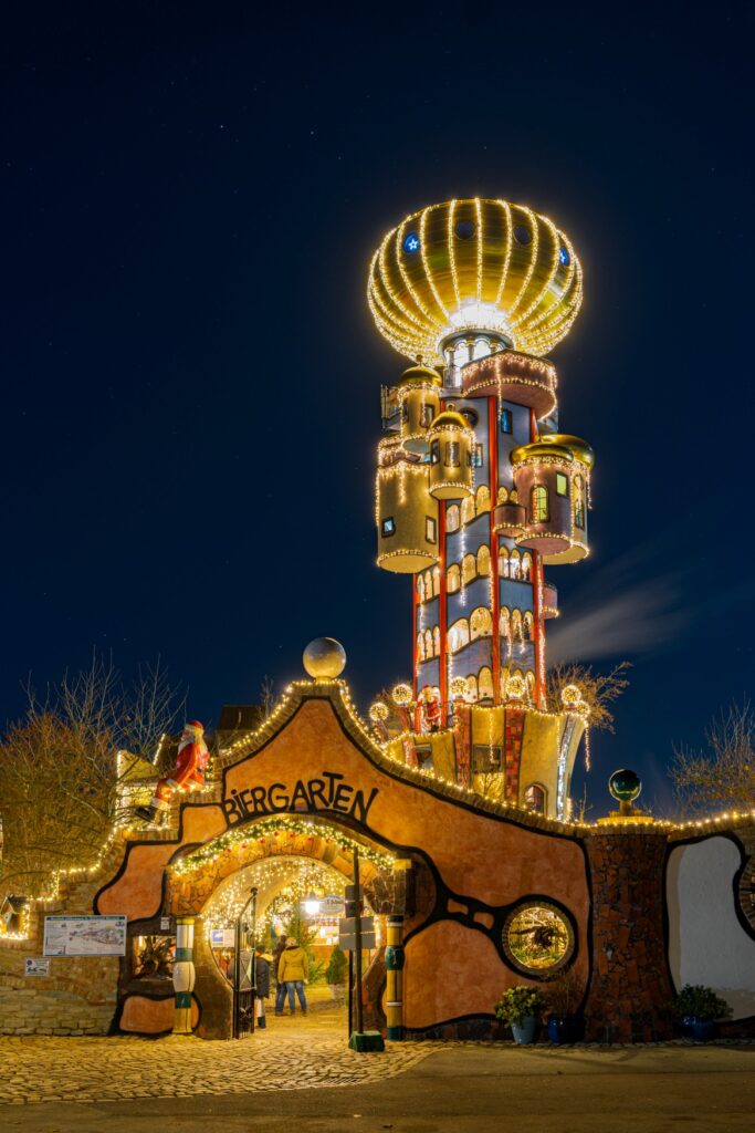 Ein hoher Turm, der wie von einem Kind gemalt aussieht, erhebt sich hinter einer braun-roten Mauer in den Nachthimmel. Auf dem Turm ist eine goldene Kuppel aufgesetzt und der ganze Turm wird von Lichterketten erleuchtet. In die Mauer vor dem Turm ist ein Tor eingelassen, über dem in handschriftlicher Optik der Schriftzug "Biergarten" angebracht ist. Auf der Mauer sitzt außerdem die Figur eines Weihnachtsmanns.