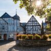 Zwei gut gepflegte Fachwerkhäuser stehen an einem gepflasterten Platz, davor ein gemauerter Brunnen mit Statue, um den kreisrund ein Blumenbeet angelegt wurde. Die Sonne scheint auf das Ensemble.